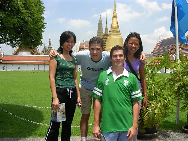 Four friends smiling in front of Wat Phra Kaew, green lawn and golden stupas