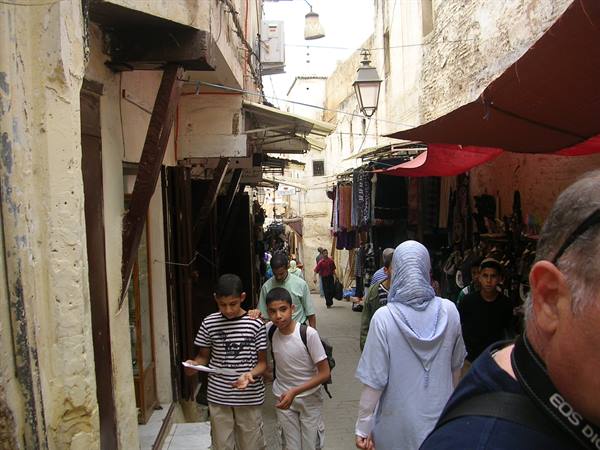 Tourists and boys walking through the Fez souk, among old walls and red awnings