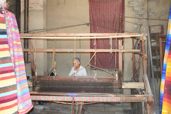 Weaver at a wooden loom among colorful carpets in a craft workshop