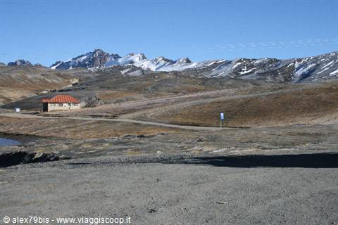 Red-roofed hut among barren hills and snow-capped mountains under blue sky
