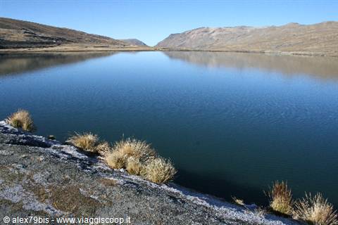 Andean lake surrounded by arid hills, tufts of yellow grass among stones