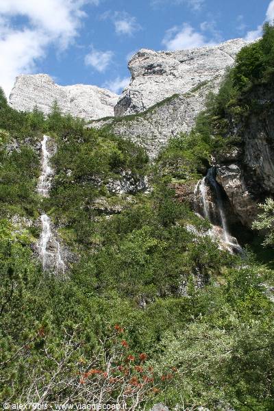 Waterfalls among larches beneath Dolomite rock walls under blue sky