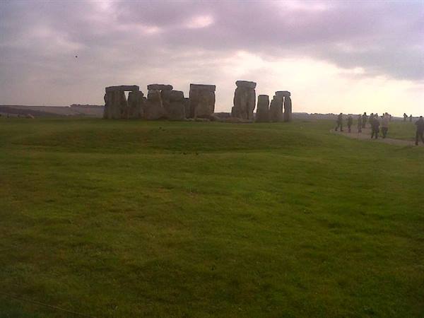 Visitors walk across green grass toward ancient Stonehenge stones under cloudy sky