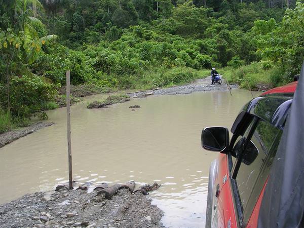Red car at muddy forest river crossing, a motorcyclist waiting on far bank