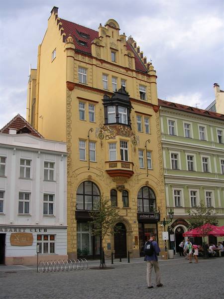 Yellow art nouveau building with black oriel window, pedestrians on cobblestone square
