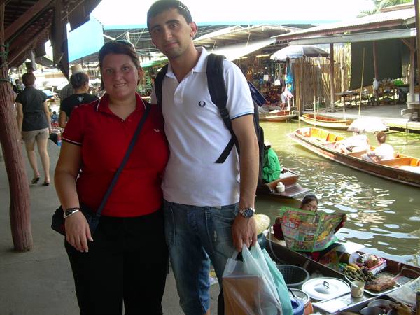 Two tourists with shopping bag at Damnoen Saduak floating market, wooden boats on sunny water