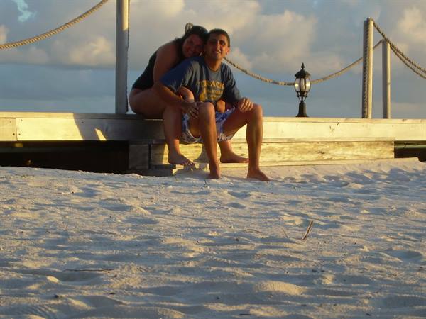 Couple sitting on wooden pier with rope railings, white sand and lantern