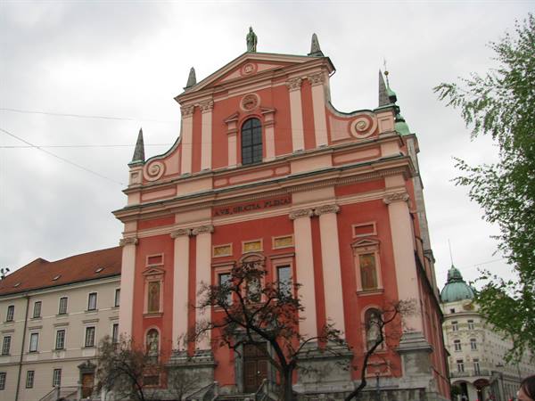 Pink Franciscan church in Ljubljana under cloudy sky, bare trees in front
