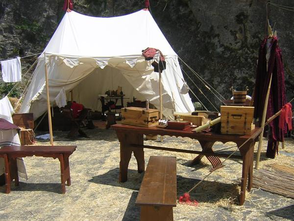 White medieval tent with wooden tables and chests, historical camp beneath rocky cliff