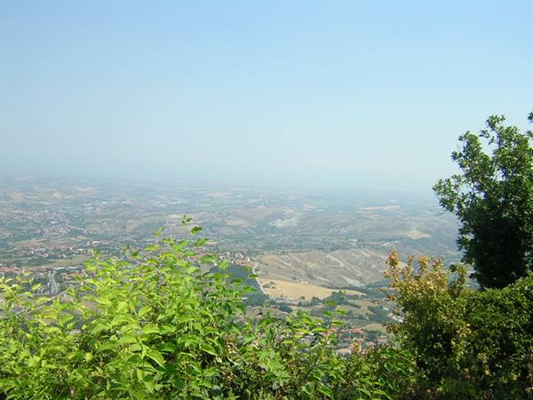 Farmed hills and small towns under hazy sky, green branches in foreground