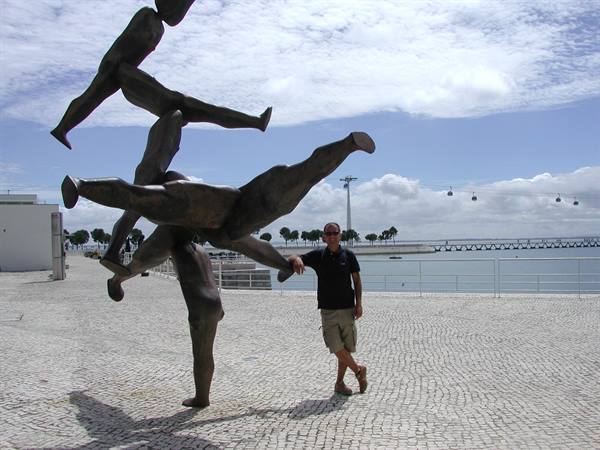 Man posing beside a bronze sculpture of figures on Lisbon's waterfront