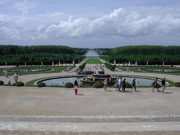 Tourists walk beside the fountain in the gardens of Versailles under white clouds