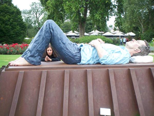 Tourist crouching behind a hyperrealistic statue of a lying man in a flowered park