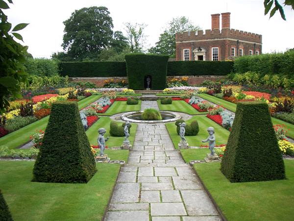 Stone path between conical hedges and statues, flowerbeds, brick palace in background