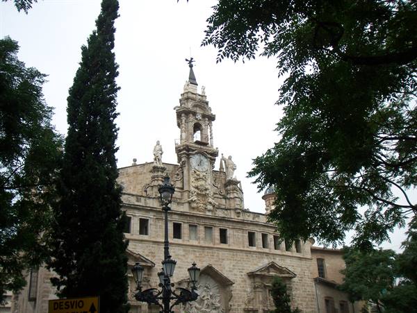 Baroque stone facade with clock and spire, framed by cypress and iron lamppost