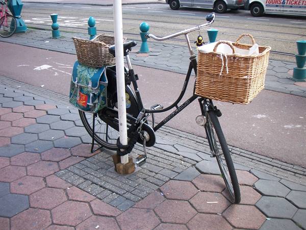 Black bicycle chained to a pole, with wicker baskets and floral bag