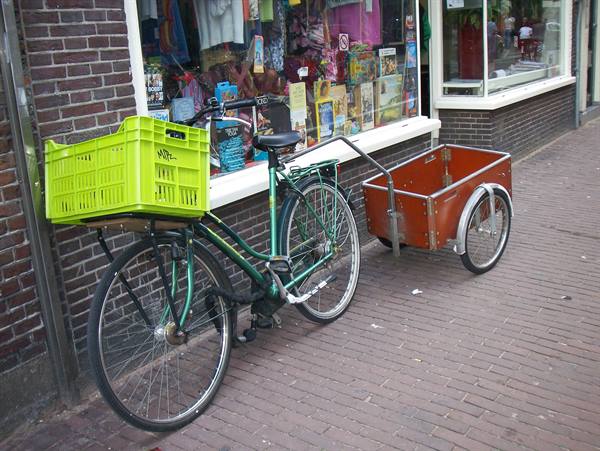 Green bicycle with green crate and red trailer parked on brick sidewalk outside a shop