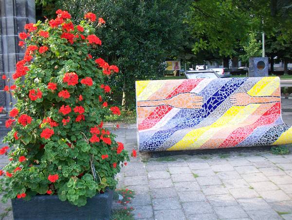 Colorful mosaic bench with intertwined arms, next to red geraniums