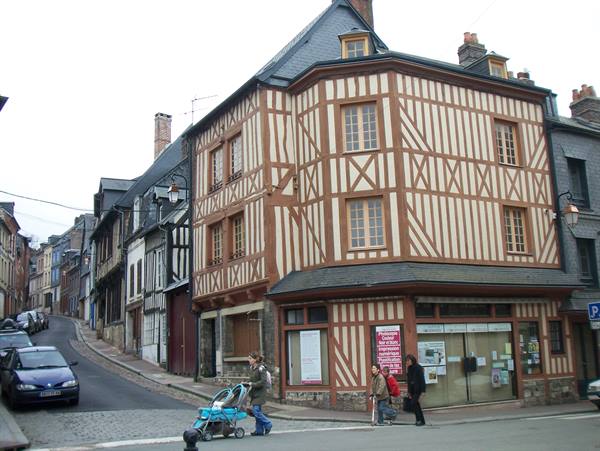 Woman with stroller crosses past a corner half-timbered house under grey sky