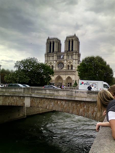 Two women on a Seine bridge gaze at Notre-Dame's Gothic facade under grey sky