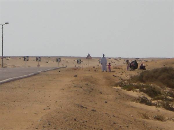Man in white and children on sand beside a desert road