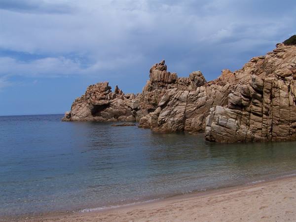 Pink granite cliffs over turquoise water, pale sandy beach, cloudy sky