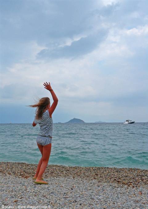 Girl in striped shirt waving from pebble beach toward distant island and boat
