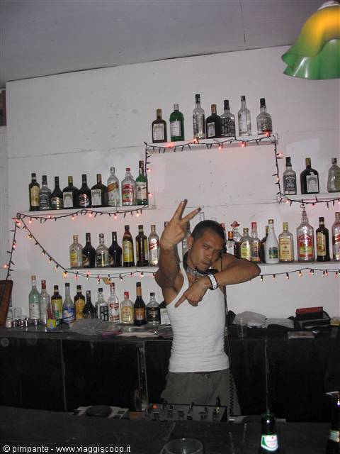 Bartender in white tank top poses with hands behind bar full of bottles