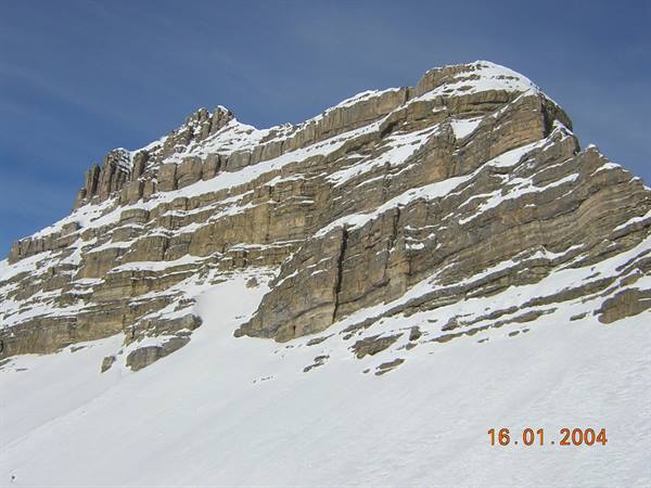 Snow-dusted layered rock face rising above a snowy slope, blue sky
