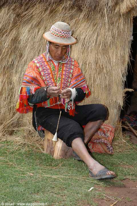 Quechua man in colorful poncho spins wool with spindle before thatched hut