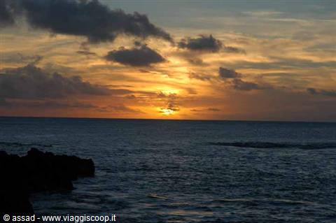 Low sun sinking into the ocean among dark clouds, rocks in silhouette