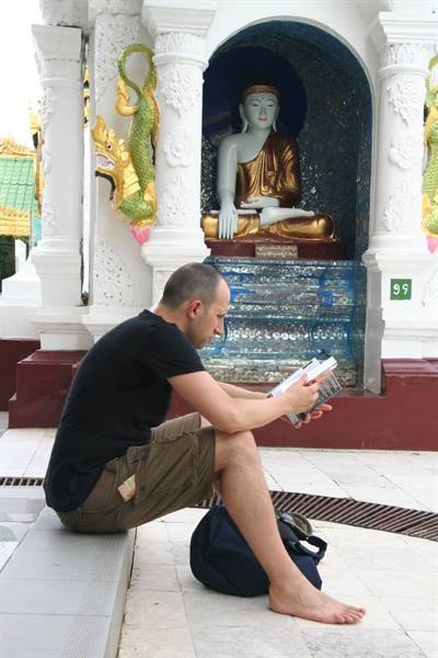 Barefoot man reads a book beside a golden Buddha statue in a pagoda