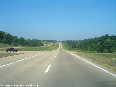 Nearly empty highway crossing green fields and trees, blue sky