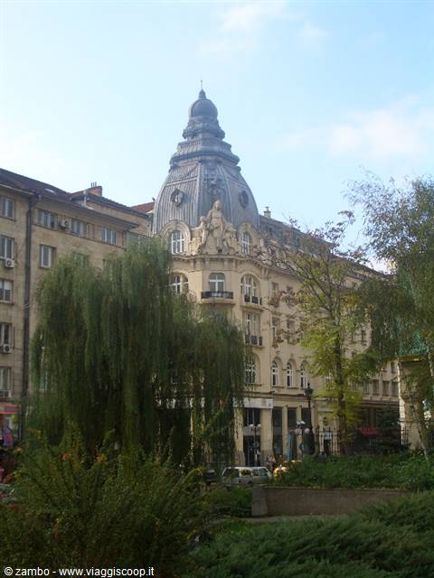Art Nouveau building with grey dome amid park trees, cars parked below