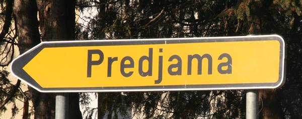 Yellow directional arrow sign pointing to Predjama, trees in background