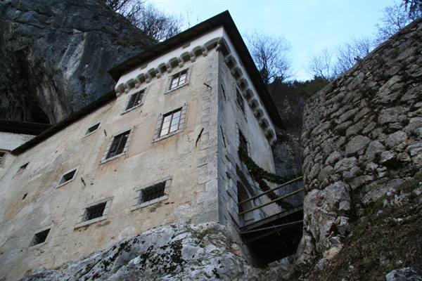 Predjama Castle: pale stone facade built into cliff rock, wooden bridge