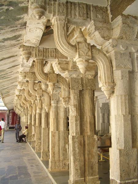 Carved stone colonnade with brackets, visitors walking in the shaded hall