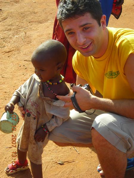 Tourist in yellow tank top smiles beside a beaded child on the red dirt