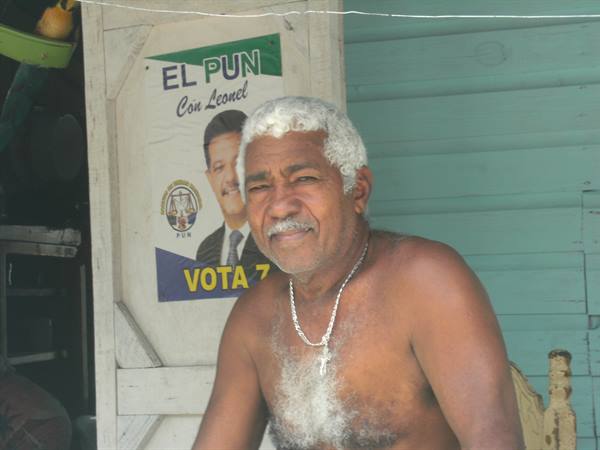 White-haired man wearing silver chain in front of an election poster