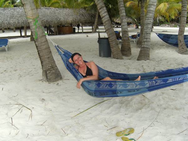 Smiling woman in black swimsuit relaxing on blue hammock between palm trees on white sand