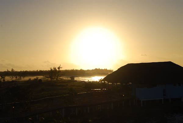 Low sun over the ocean, thatched-roof bungalow silhouetted on the beach