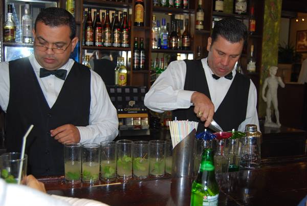 Two bartenders in black vests prepare mojitos with mint on a wooden counter