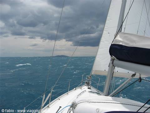 White sail over choppy turquoise sea, dark clouds and distant island