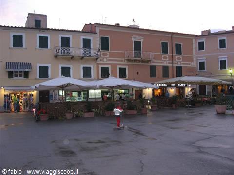 Child on scooter in a small square with umbrellas and pastel buildings, Marciana Marina