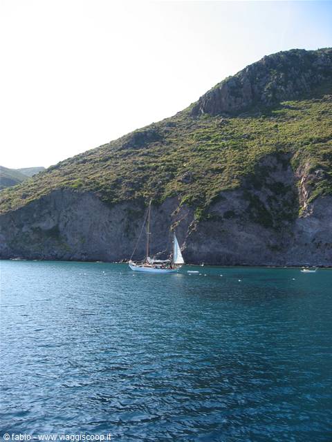 White sailboat anchored before green cliffs covered in Mediterranean scrub
