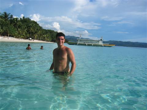 Man wading in turquoise water near white beach with boat and palm trees