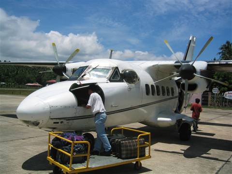 Man loads luggage onto white twin-engine turboprop on tropical airstrip