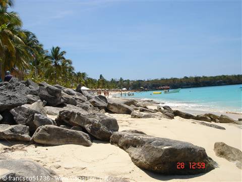 Rocks on white sand in front of turquoise sea with boats and palms