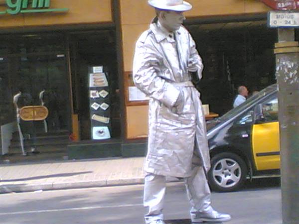 Silver mime in hat and trench coat standing still beside a yellow taxi