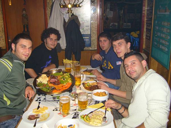 Five friends at a tavern table with grilled meat platters and beer mugs
